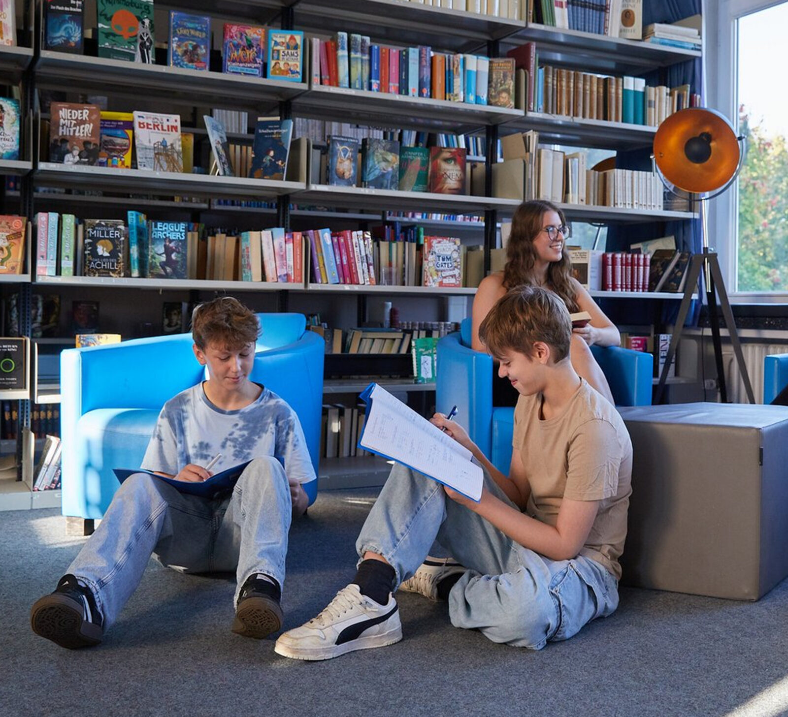 Drei Jugendliche lernen in einer Bibliothek. Zwei sitzen auf dem Boden und schreiben in ihre Hefte, w&auml;hrend einer auf einem Stuhl sitzt und liest. Im Hintergrund sind B&uuml;cherregale, bunte B&uuml;cher und eine Stehlampe zu sehen.