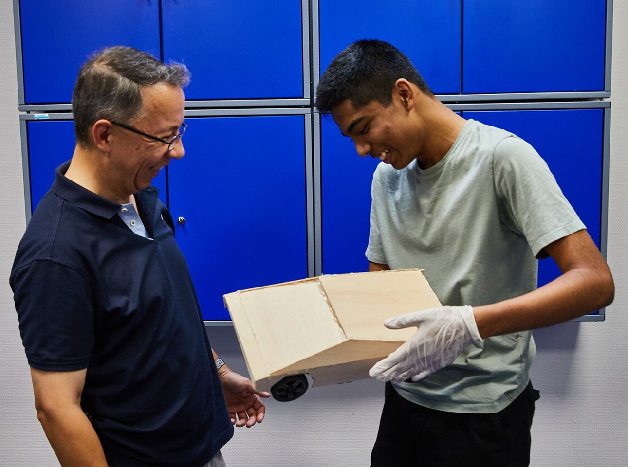 Zwei Personen lächeln, während sie ein kleines Modellauto aus Holz betrachten. Eine Person, die Handschuhe trägt, hält das Auto in der Hand, während die andere zusieht. Im Hintergrund sind blaue Spinde zu sehen.