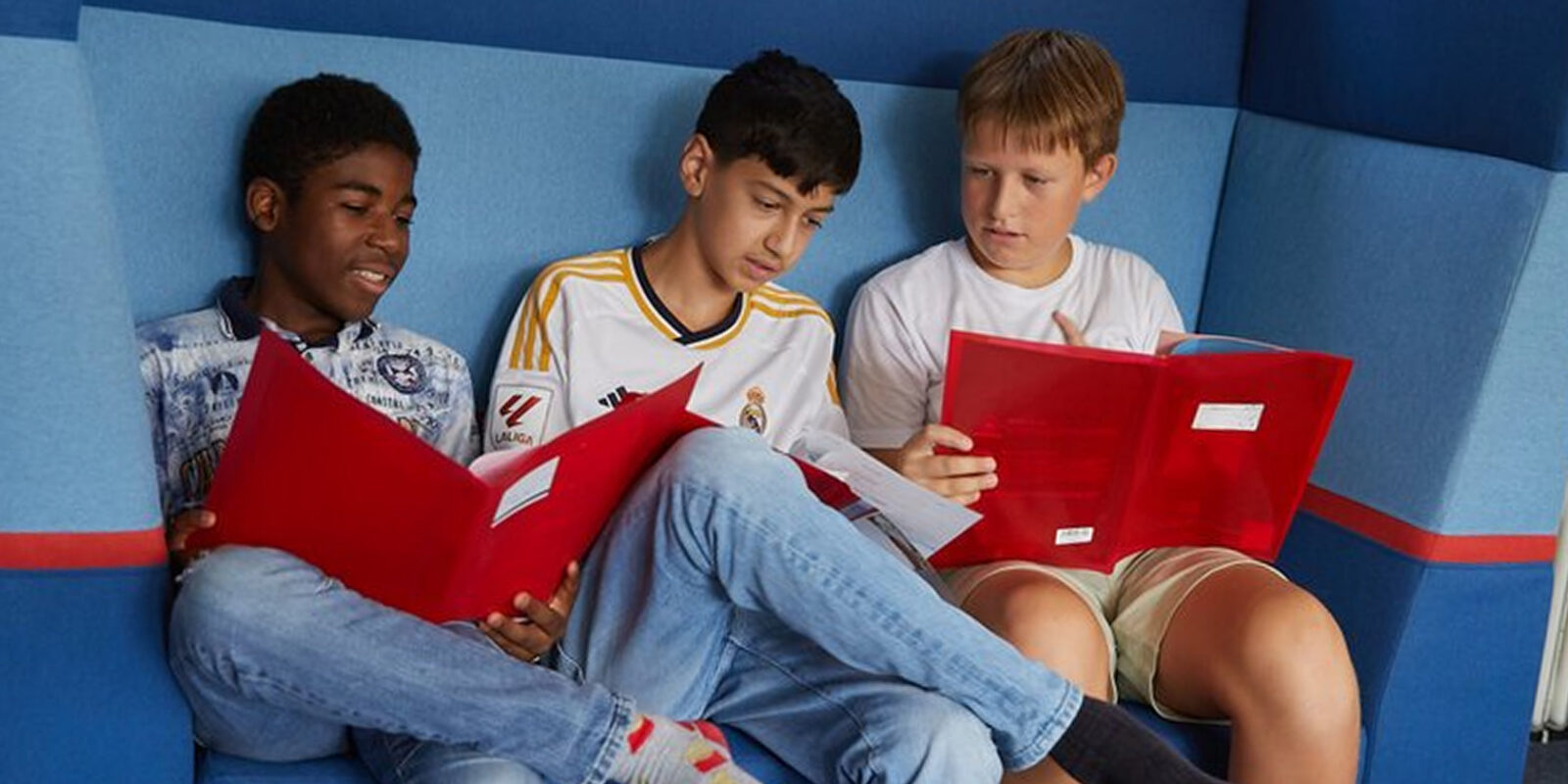 Drei Jungen sitzen zusammen auf einem blauen Sofa, jeder hält eine rote Mappe in der Hand und schaut auf seine Unterlagen; es sieht so aus, als würden sie gemeinsam lernen oder lesen.