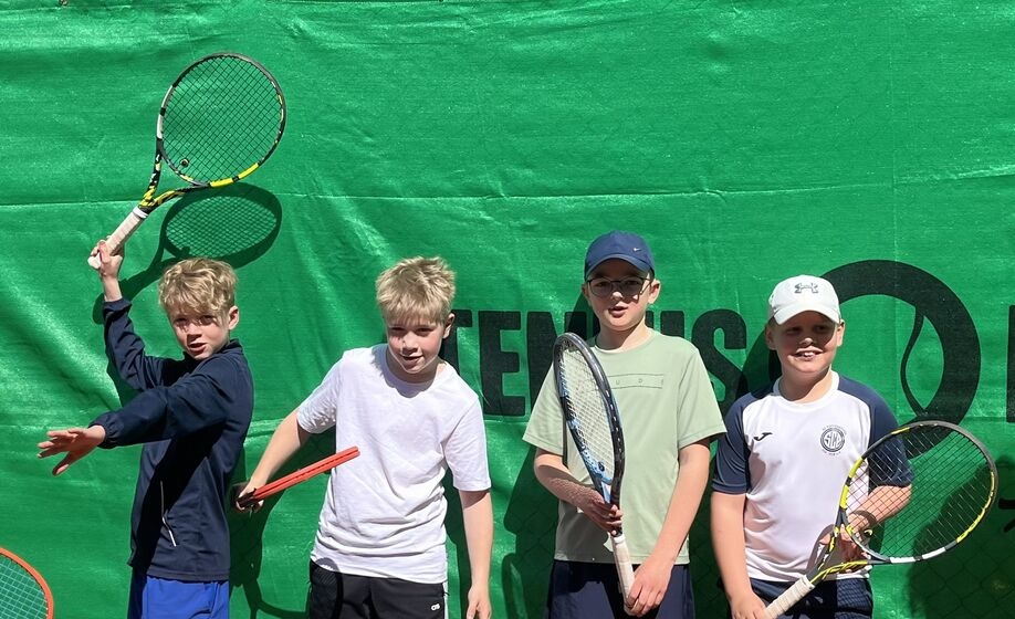 Vier Jungen stehen vor einer grünen Tennisplatzkulisse und halten jeweils einen Tennisschläger in der Hand. Sie tragen Sportkleidung und scheinen an einem sonnigen Tag für ein Foto zu posieren.
