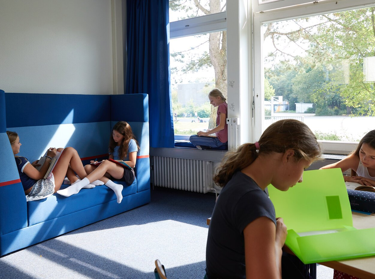 Vier Mädchen lesen und lernen still in einem hellen Klassenzimmer. Zwei sitzen auf einem blauen Sofa, eines auf der Fensterbank und ein weiteres an einem Schreibtisch mit einer Mappe. Sonnenlicht strömt durch die großen Fenster herein.