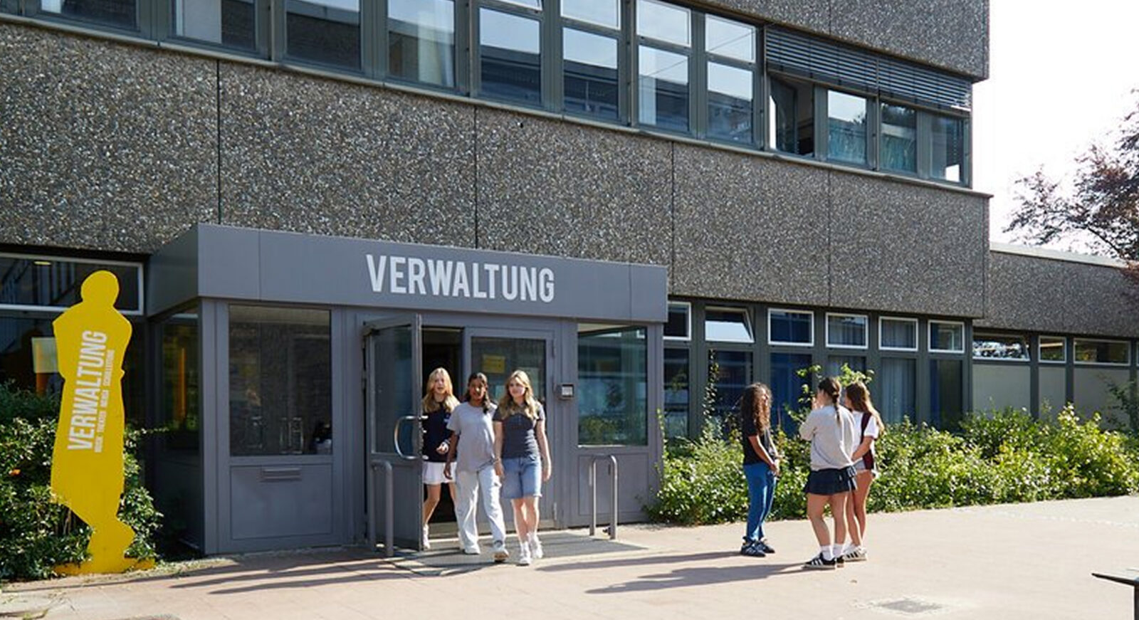 Eine Gruppe von Schülern steht vor einem modernen Schulgebäude, über dessen Eingang das Schild „Verwaltung“ prangt, und geht daran vorbei. In der Nähe des Eingangs befindet sich eine gelbe Silhouette, auf der „Verwaltung“ steht.