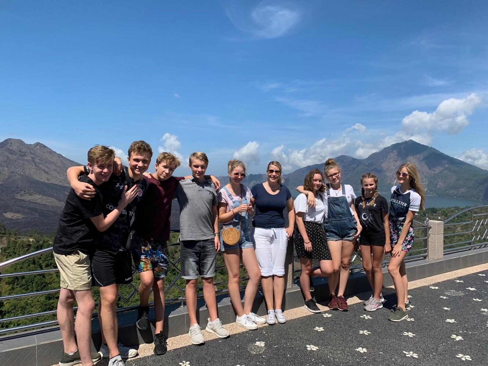 Eine Gruppe von zehn Jugendlichen und einem Erwachsenen steht an einem sonnigen Tag lächelnd im Freien, im Hintergrund sind Berge und blauer Himmel zu sehen. Sie befinden sich an einem Aussichtspunkt mit Geländer und gemustertem Pflaster.