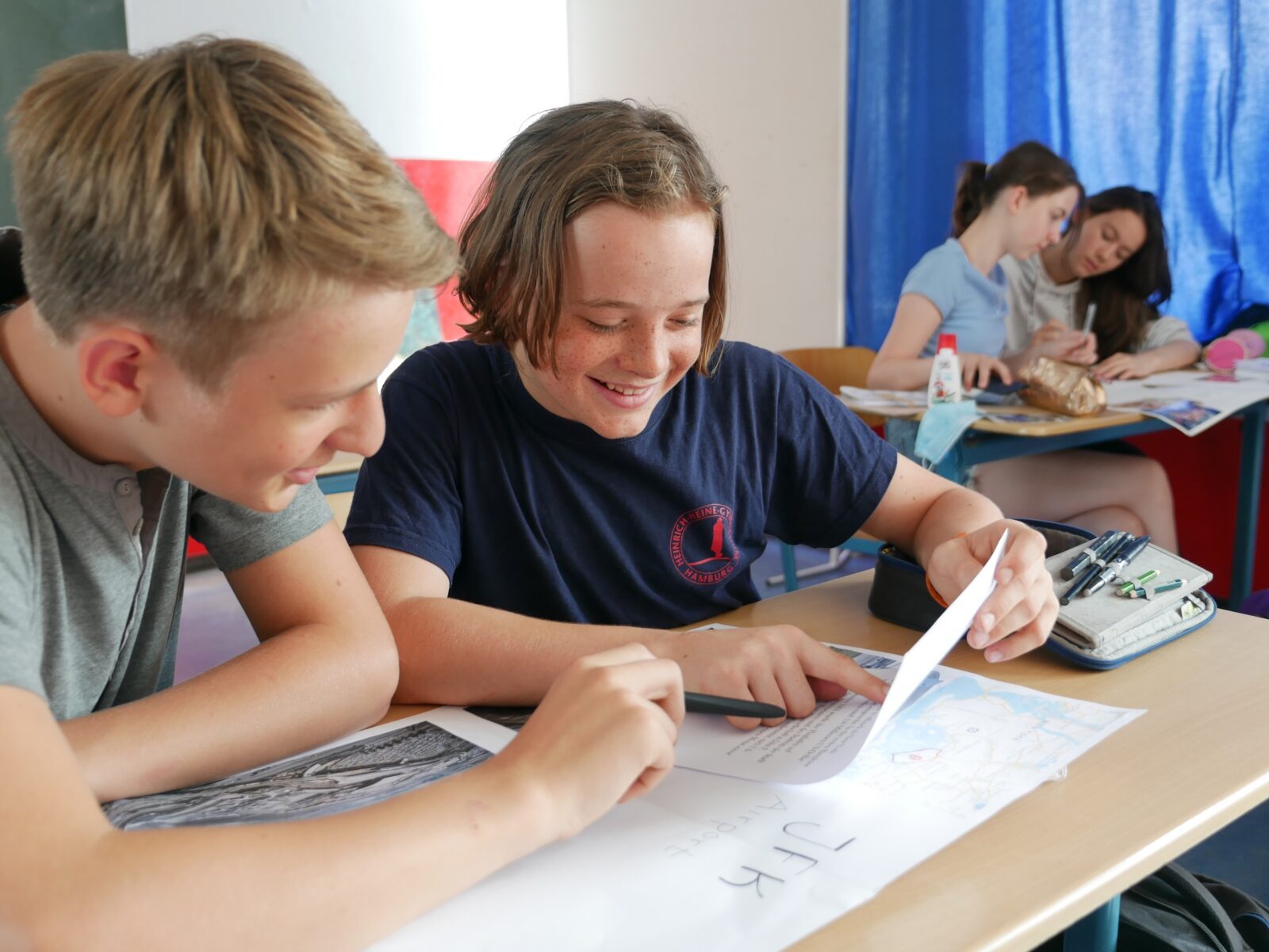 Zwei Schüler sitzen an einem Pult, lächeln und schauen gemeinsam auf ein Blatt Papier, während im Hintergrund zwei weitere Schüler an einem anderen Pult in einem Klassenzimmer arbeiten.