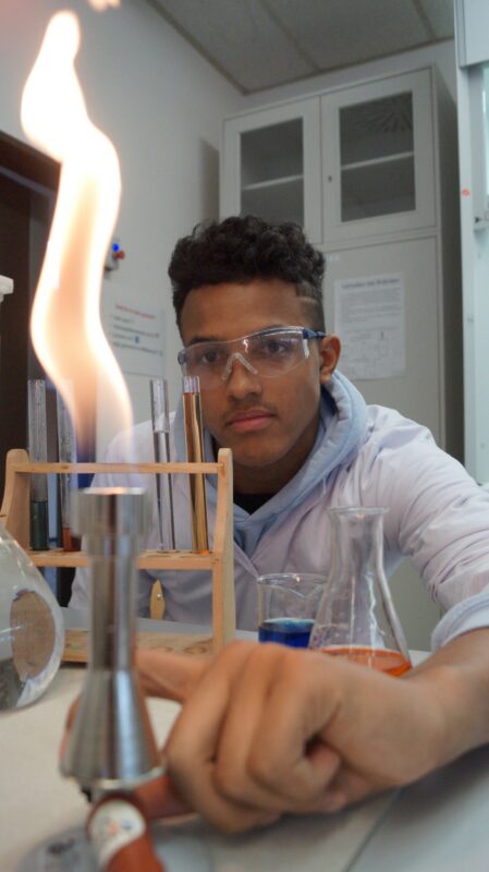 Ein Schüler, der eine Schutzbrille und einen Laborkittel trägt, beobachtet in einem Chemielabor eine hohe orangefarbene Flamme eines Bunsenbrenners, während in der Nähe Reagenzgläser und Kolben mit bunten Flüssigkeiten stehen.