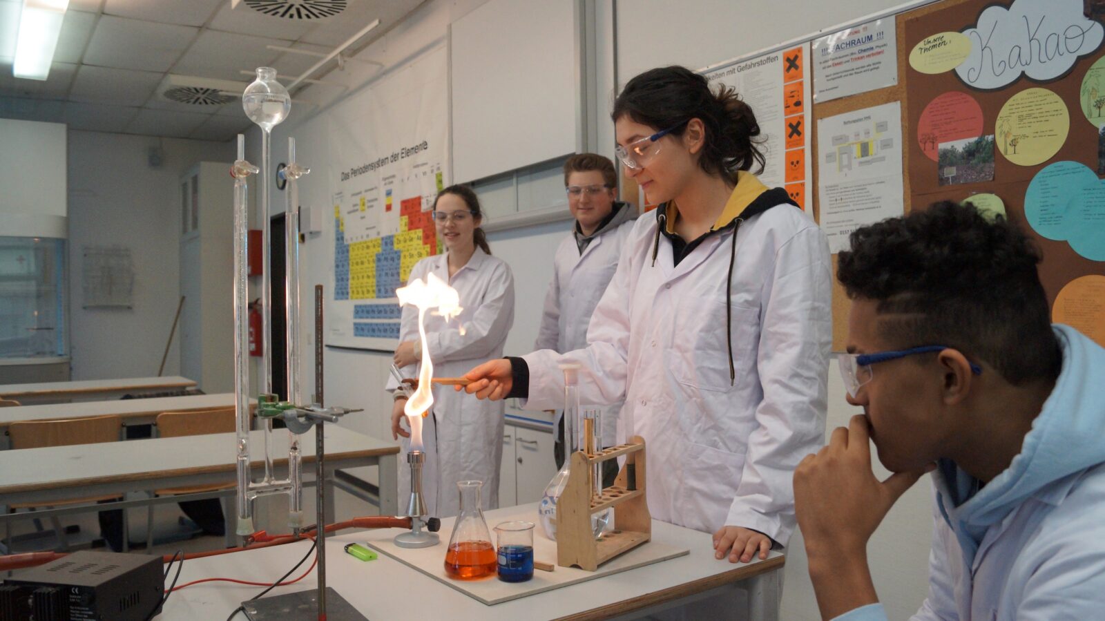Vier Schüler in weißen Laborkitteln führen in einem Klassenzimmer ein Chemieexperiment durch. Ein Schüler hält ein Werkzeug über eine Flamme, während auf dem Tisch bunte Flüssigkeiten und Laborgeräte stehen. Die anderen Schüler beobachten das Geschehen aufmerksam.