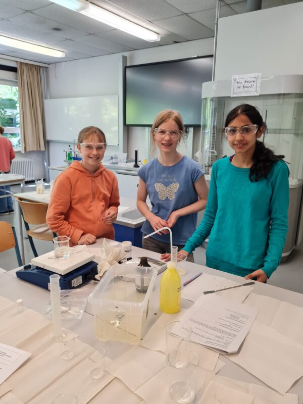 Drei Mädchen mit Schutzbrillen stehen an einem Labortisch, auf dem Bechergläser, ein Messzylinder, Papiere und andere Laborutensilien liegen, und lächeln in einem hellen Naturwissenschaftsraum in die Kamera.