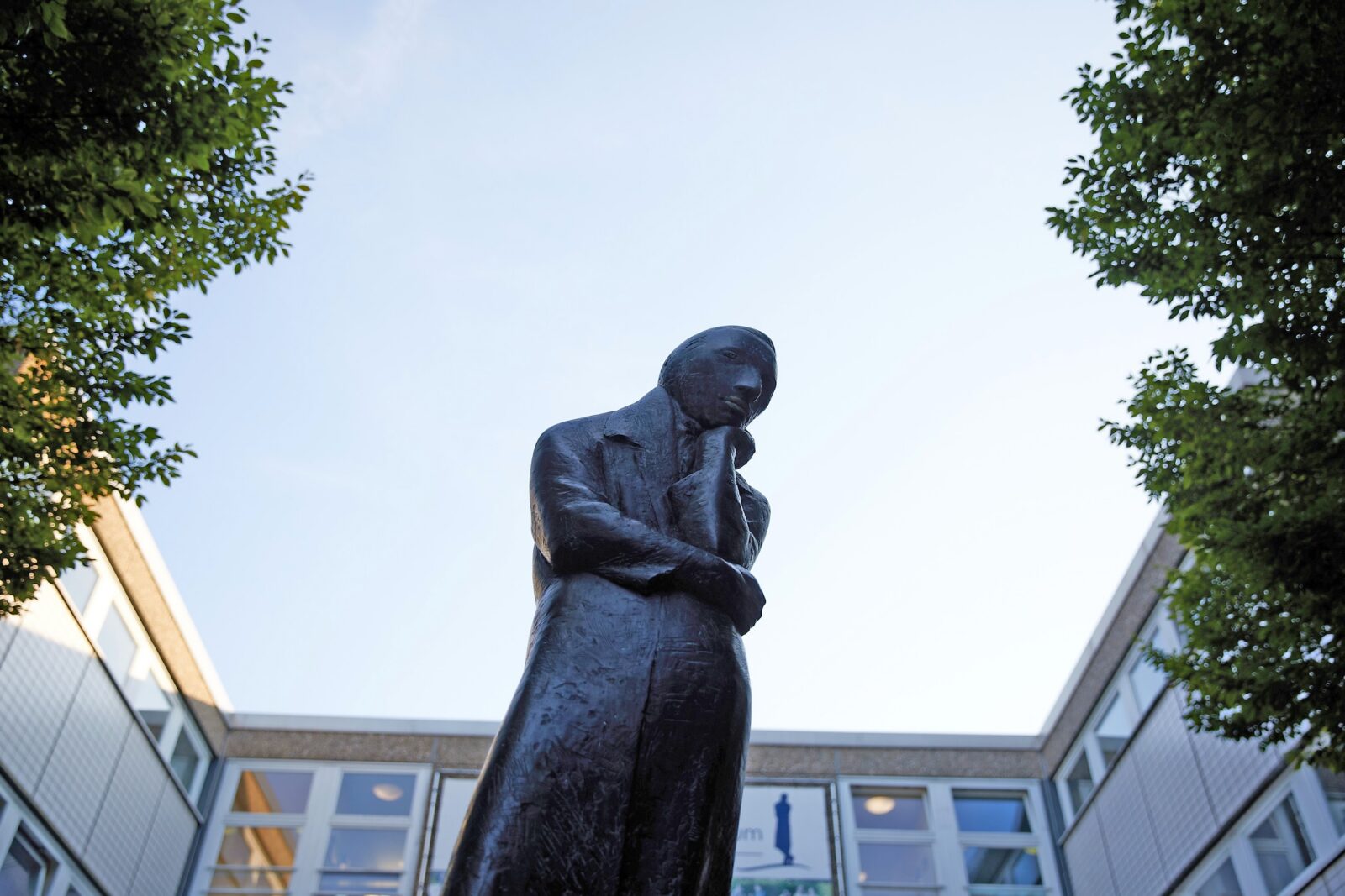 Eine Bronzestatue einer stehenden Person, die eine Hand am Kinn hat, umgeben von Gebäuden und Bäumen, von unten betrachtet vor einem klaren Himmel.