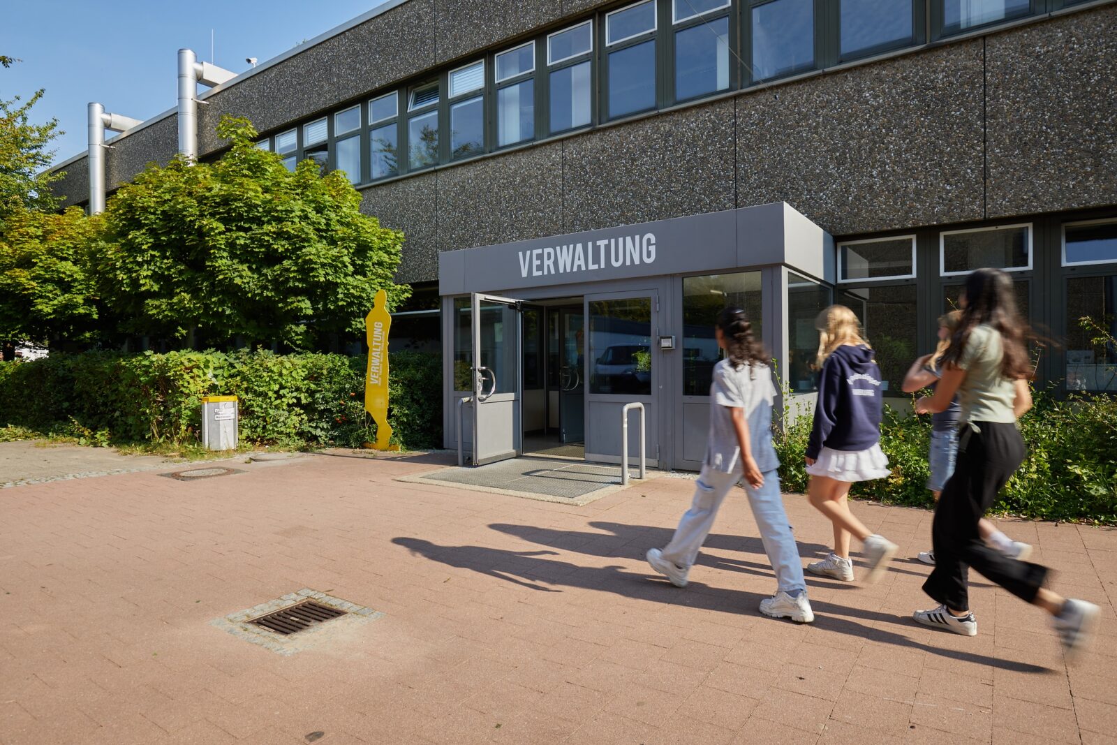 An einem sonnigen Tag gehen drei junge Leute am Eingang eines Gebäudes vorbei, auf dem „Verwaltung“ steht. Das Gebäude hat große Fenster und ist von Grünflächen umgeben.