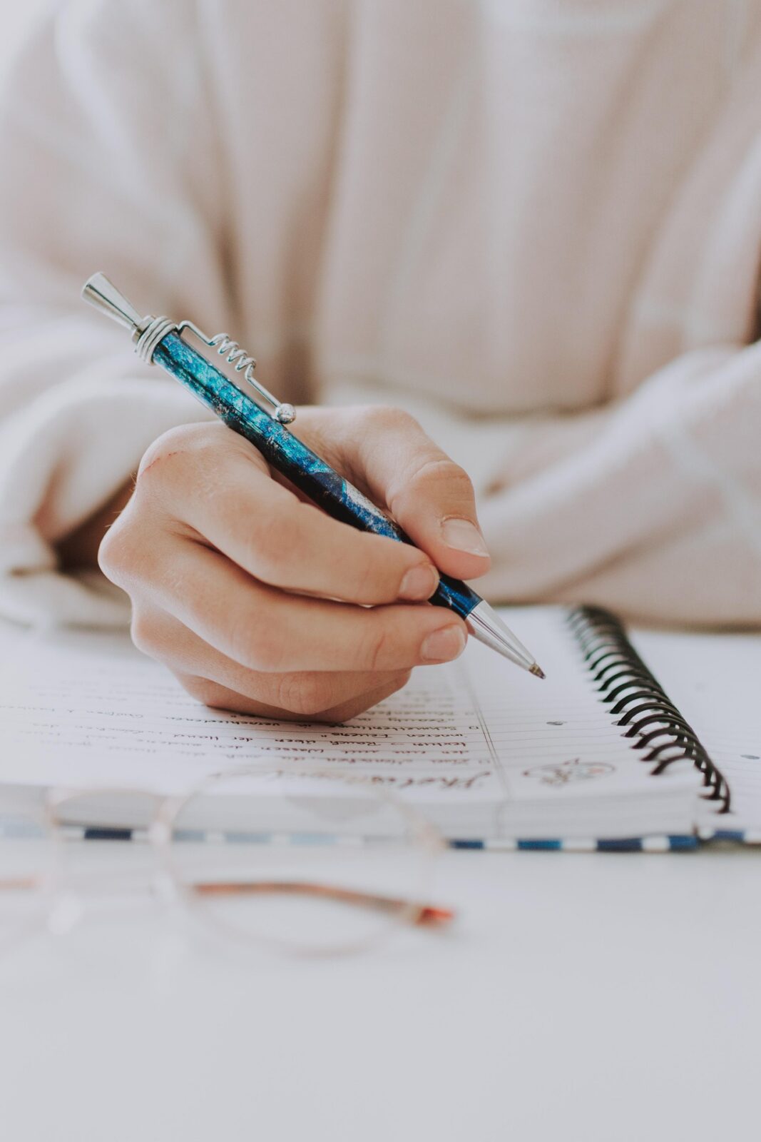 Eine Person, die einen leichten Pullover trägt, hält einen blauen Stift in der Hand und schreibt in ein Spiralheft, während ihre Brille auf dem weißen Tisch im Vordergrund liegt.