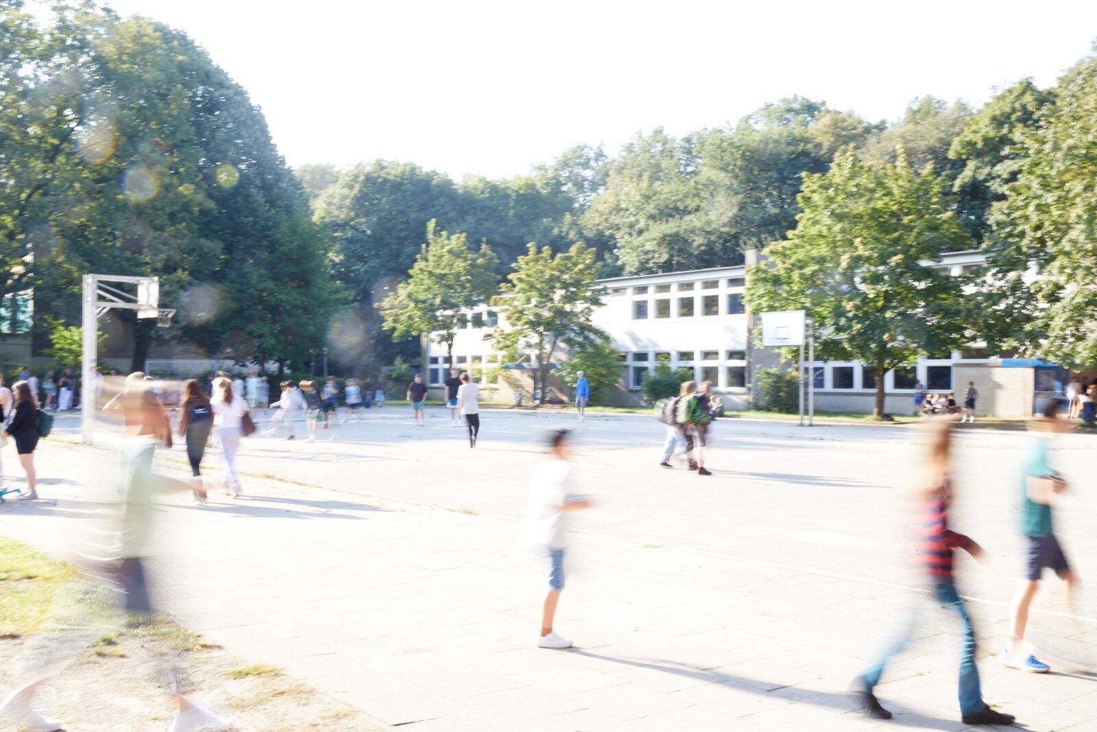 Ein strahlender, sonniger Tag auf einem Schulhof, auf dem viele Kinder herumlaufen und spielen. Im Hintergrund ist ein weißes Schulgebäude zu sehen, das von Bäumen umgeben ist. Das Bild ist leicht überbelichtet und unscharf, was einen Eindruck von Bewegung vermittelt.