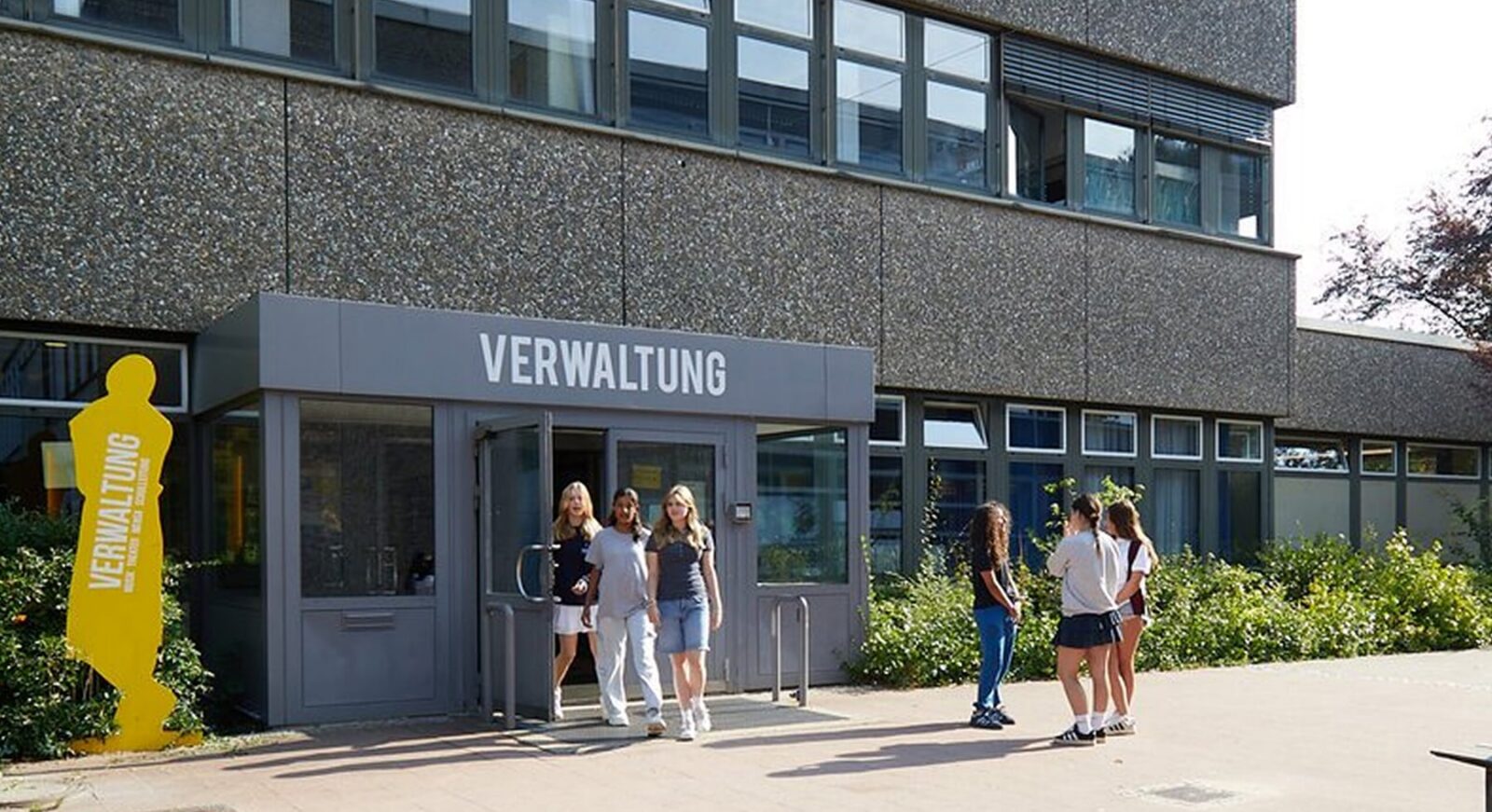 Mehrere Schüler gehen an einem modernen Schulgebäude mit großen Fenstern vorbei. Am Eingang befindet sich ein Schild mit der Aufschrift „VERWALTUNG“, und neben der Tür steht eine gelbe Silhouette, umgeben von Grünpflanzen.