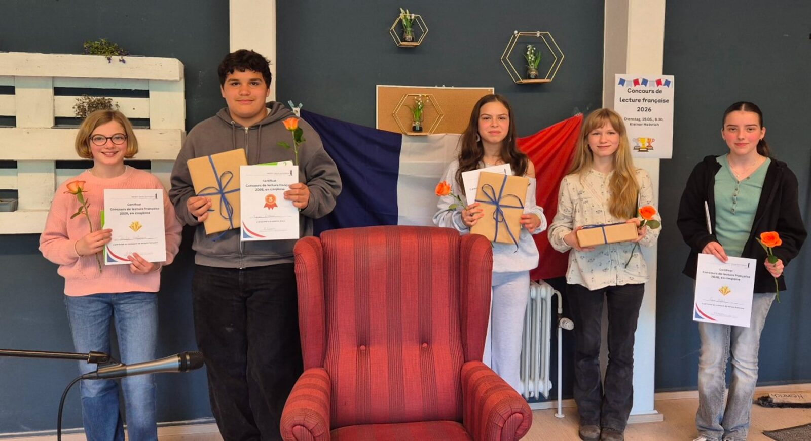 Fünf lächelnde Teenager stehen in einem Raum und halten Urkunden, orangefarbene Blumen und verpackte Geschenke in den Händen. Hinter ihnen stehen ein roter Sessel, eine französische Flagge und eine Wand mit Regalen und gerahmten Gegenständen.