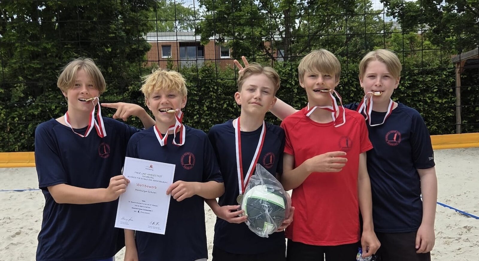 Fünf Jungen stehen auf einem sandigen Volleyballfeld, tragen Medaillen um den Hals und lächeln. Einer hält eine Urkunde in der Hand, ein anderer einen Preis. Sie wirken glücklich und feiern einen Sieg. Im Hintergrund sind Bäume und ein Netz zu sehen.