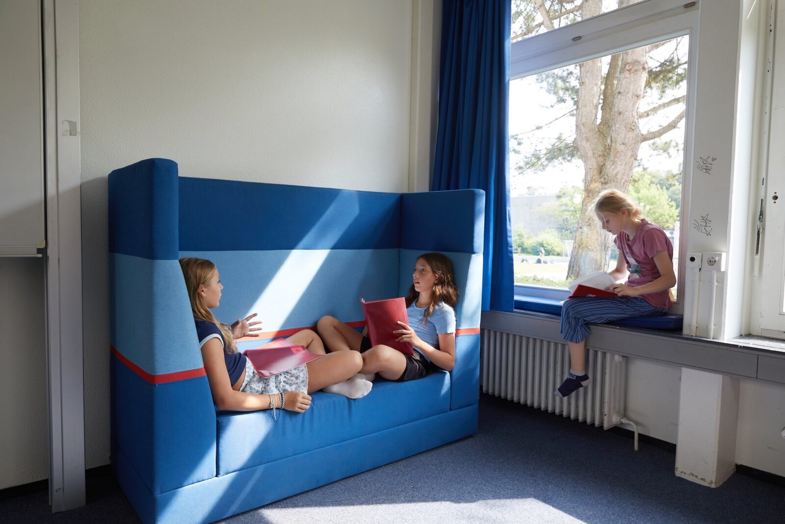 Drei Mädchen befinden sich in einem hellen Klassenzimmer. Zwei sitzen auf einem blauen, gepolsterten Sofa und unterhalten sich, während sie Ordner auf dem Schoß haben; das dritte Mädchen sitzt auf der Fensterbank und liest oder schreibt mit einem Ordner in der Hand.