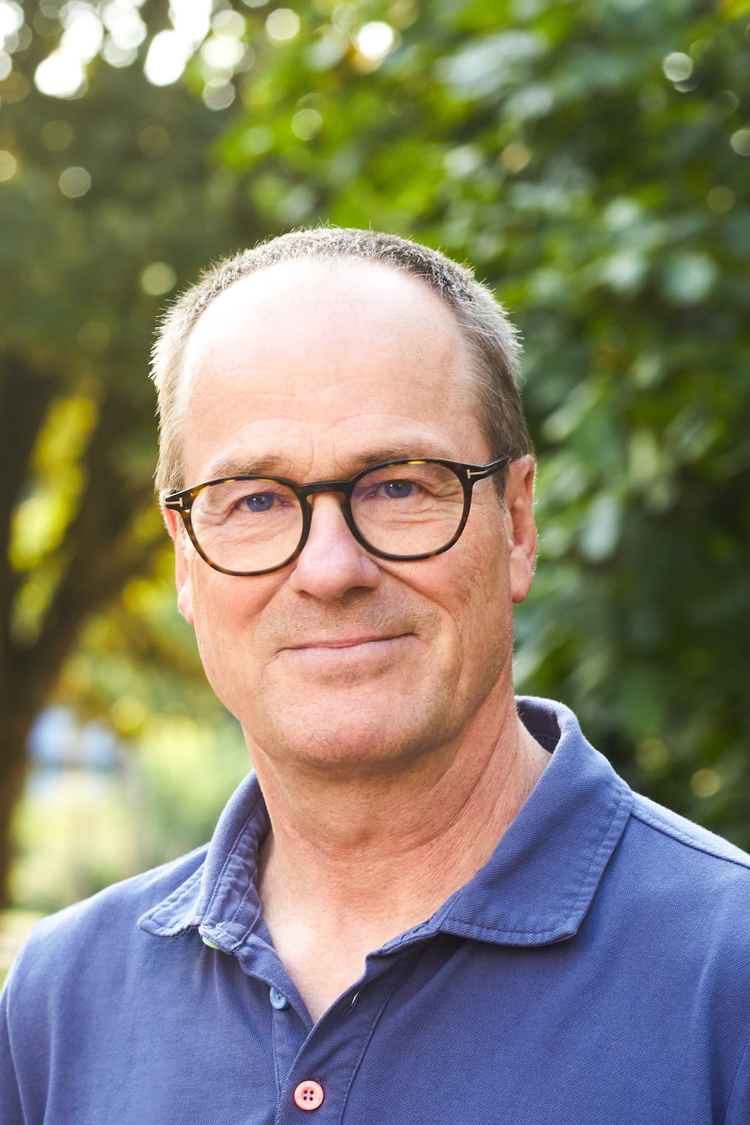 Ein Mann mittleren Alters mit kurzen Haaren und Brille, der ein blaues Hemd mit Kragen trägt, steht draußen vor grünem Laub und im Sonnenlicht.