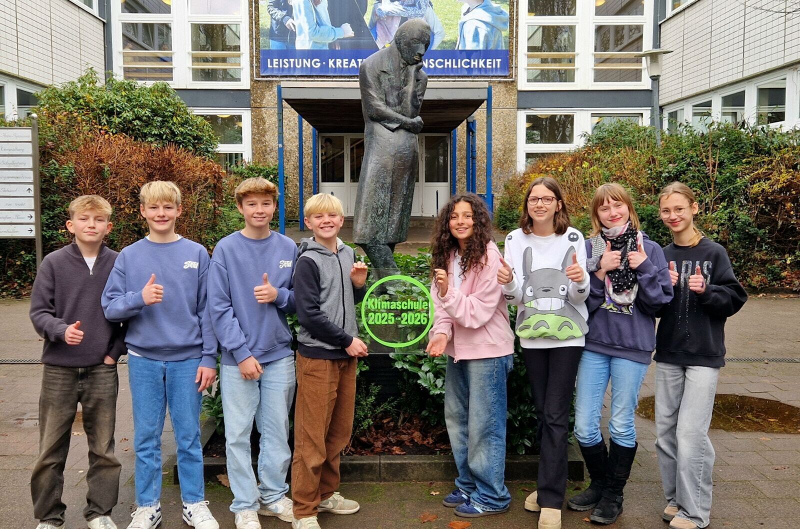 Acht lächelnde Teenager stehen vor einer Statue und einem Schulgebäude und zeigen den Daumen nach oben. Sie halten ein grünes Schild mit der Aufschrift „Klimaschule 2025–2026“ in den Händen. Im Hintergrund sind Büsche und ein Schild zu sehen.