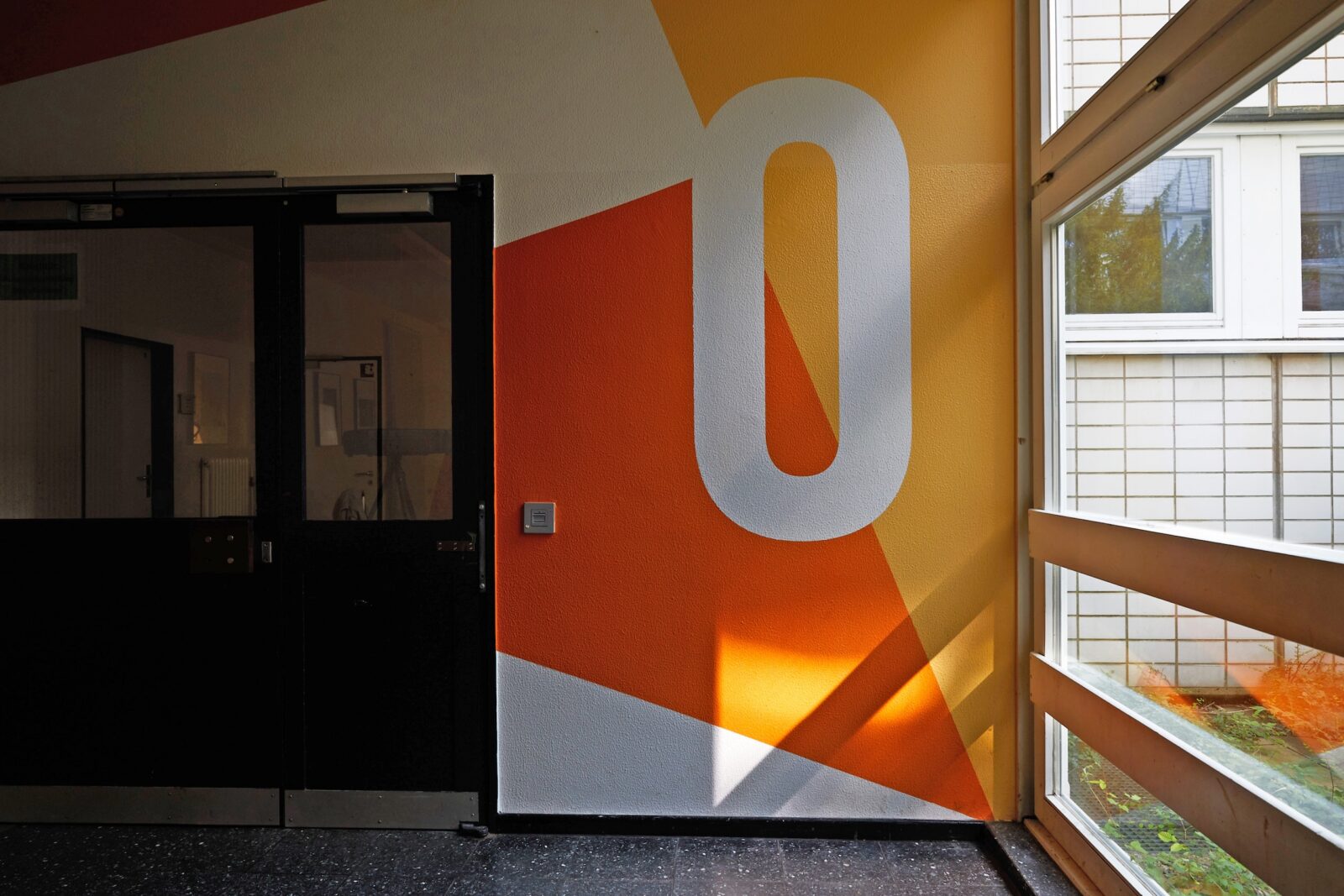 Ein Flur mit einer großen weißen 0 an einer orange-gelben Wand, neben einer schwarzen Doppeltür und großen Fenstern, durch die Sonnenlicht hereinströmt. Der Boden ist dunkel und gesprenkelt.