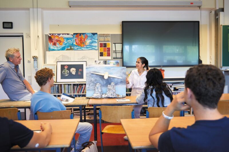 Ein Schüler präsentiert einer Gruppe im Klassenzimmer seine Kunstwerke. Mehrere Schüler und ein Lehrer schauen zu, während der Vortragende auf Gemälde deutet, die auf einer Staffelei ausgestellt sind; im Raum sind Zeichnungen und Malutensilien zu sehen.