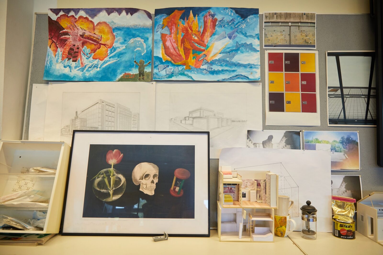 An einer Wand hängen farbenfrohe Gemälde, architektonische Skizzen und ein gerahmtes Stillleben mit einem Schädel, einer Tulpe und einer Sanduhr. Darunter befinden sich ein Miniaturmodell eines Raums, ein kleiner Stuhl und ein Glas, umgeben von Malutensilien und Papieren.
