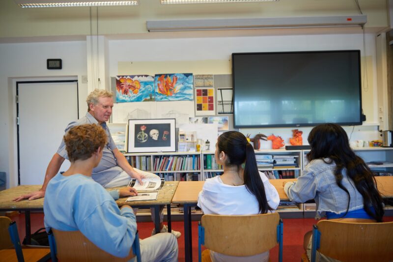 Ein Lehrer sitzt auf einem Pult vorne im Klassenzimmer und spricht mit drei Schülern, die ihm gegenüber sitzen. Im Klassenzimmer befinden sich Malutensilien, Gemälde und eine große Leinwand an der Wand.