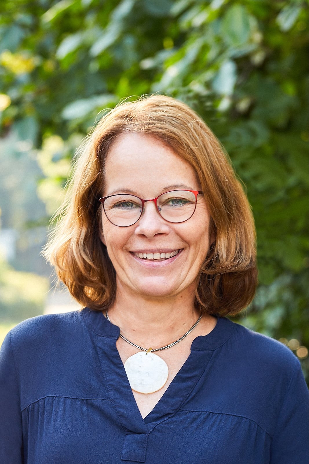 Eine Frau mit schulterlangem hellbraunem Haar, die eine Brille, eine marineblaue Bluse und eine Halskette mit einem großen runden Anhänger trägt, lächelt im Freien vor dem Hintergrund von grünem Laub.