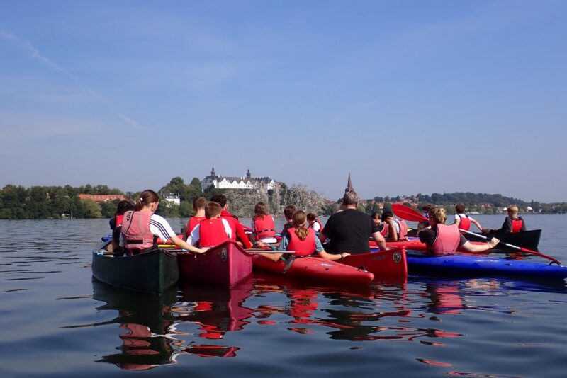 Eine Gruppe von Menschen in Schwimmwesten paddelt mit Kanus auf einem ruhigen See, vor ihnen liegen in der Ferne ein weißes Schloss und Gebäude am Ufer unter einem strahlend blauen Himmel.