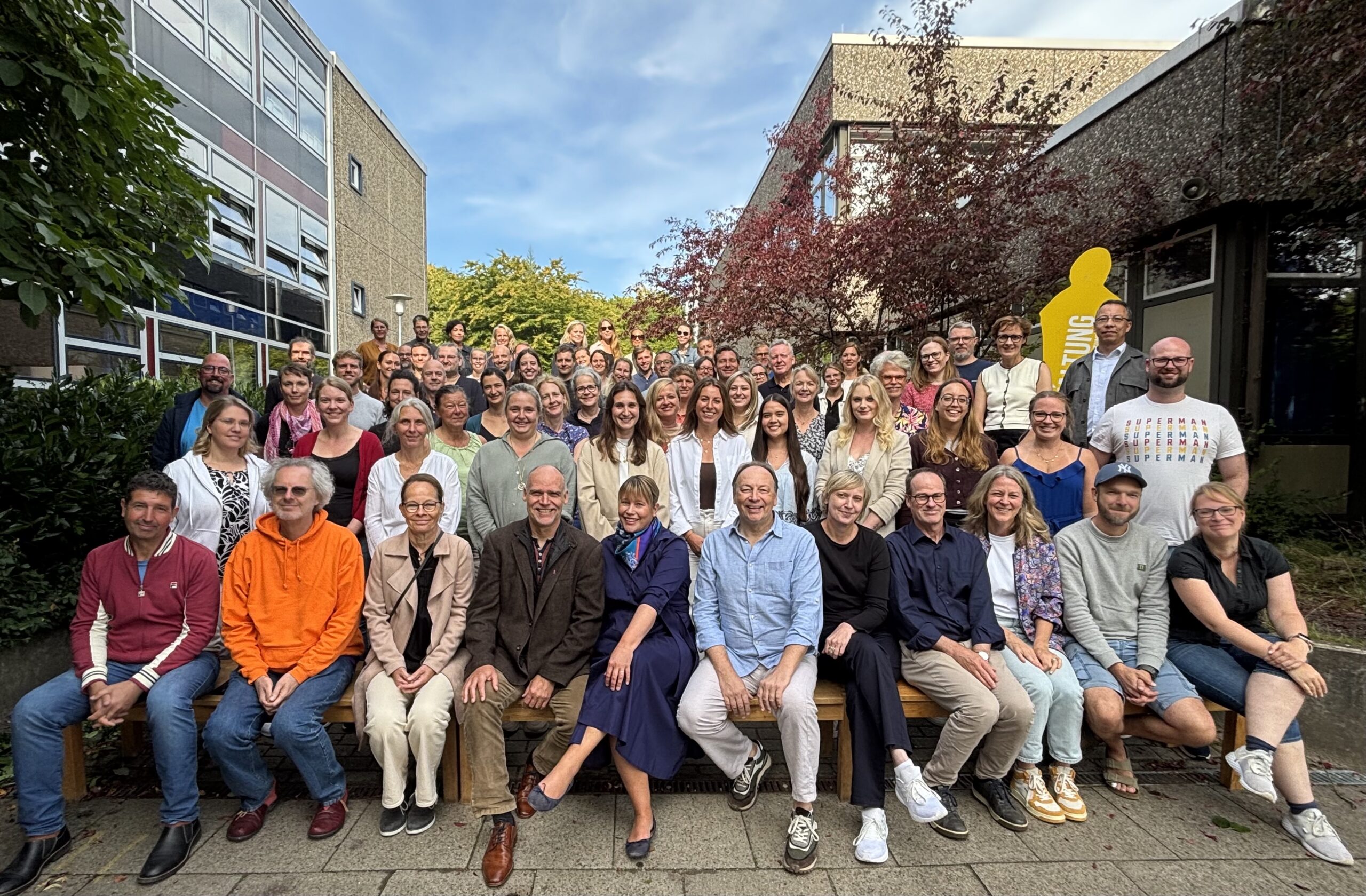 Eine große Gruppe von Menschen, die lächeln und vor einem Gebäude mit Bäumen und blauem Himmel im Hintergrund für ein Gruppenfoto posieren. Einige sitzen auf Bänken im Vordergrund, während andere hinter ihnen stehen.