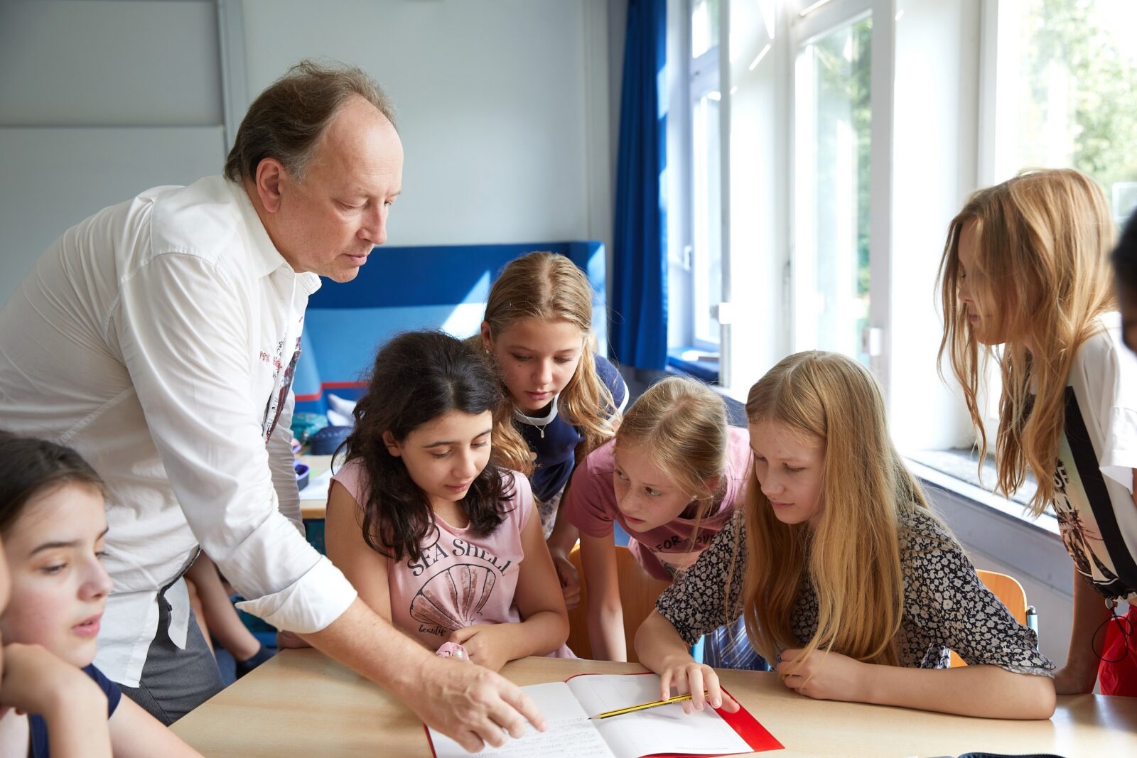 Ein Lehrer und eine Gruppe junger Schüler sitzen in einem Klassenzimmer um einen Tisch herum und schauen gemeinsam in ein aufgeschlagenes Buch, während Sonnenlicht durch die großen Fenster hereinströmt.