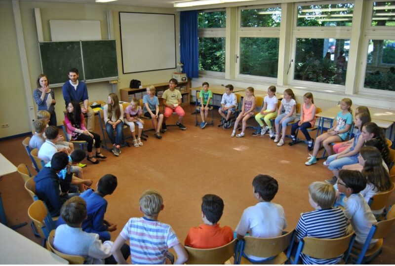 Eine Gruppe von Schulkindern sitzt in einem großen Stuhlkreis in einem Klassenzimmer, während zwei Erwachsene vorne neben einer Tafel stehen. Der Raum hat große Fenster, und die Schüler scheinen gerade an einer Gruppenaktivität teilzunehmen.