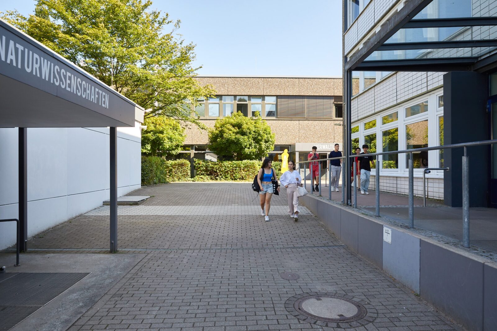An einem sonnigen Tag gehen mehrere Menschen vor einem Schul- oder Universitätsgebäude vorbei. Auf einem Schild steht „Naturwissenschaften“. Der Bereich ist mit Ziegelsteinen gepflastert, und entlang des Weges stehen Bäume und Sträucher.