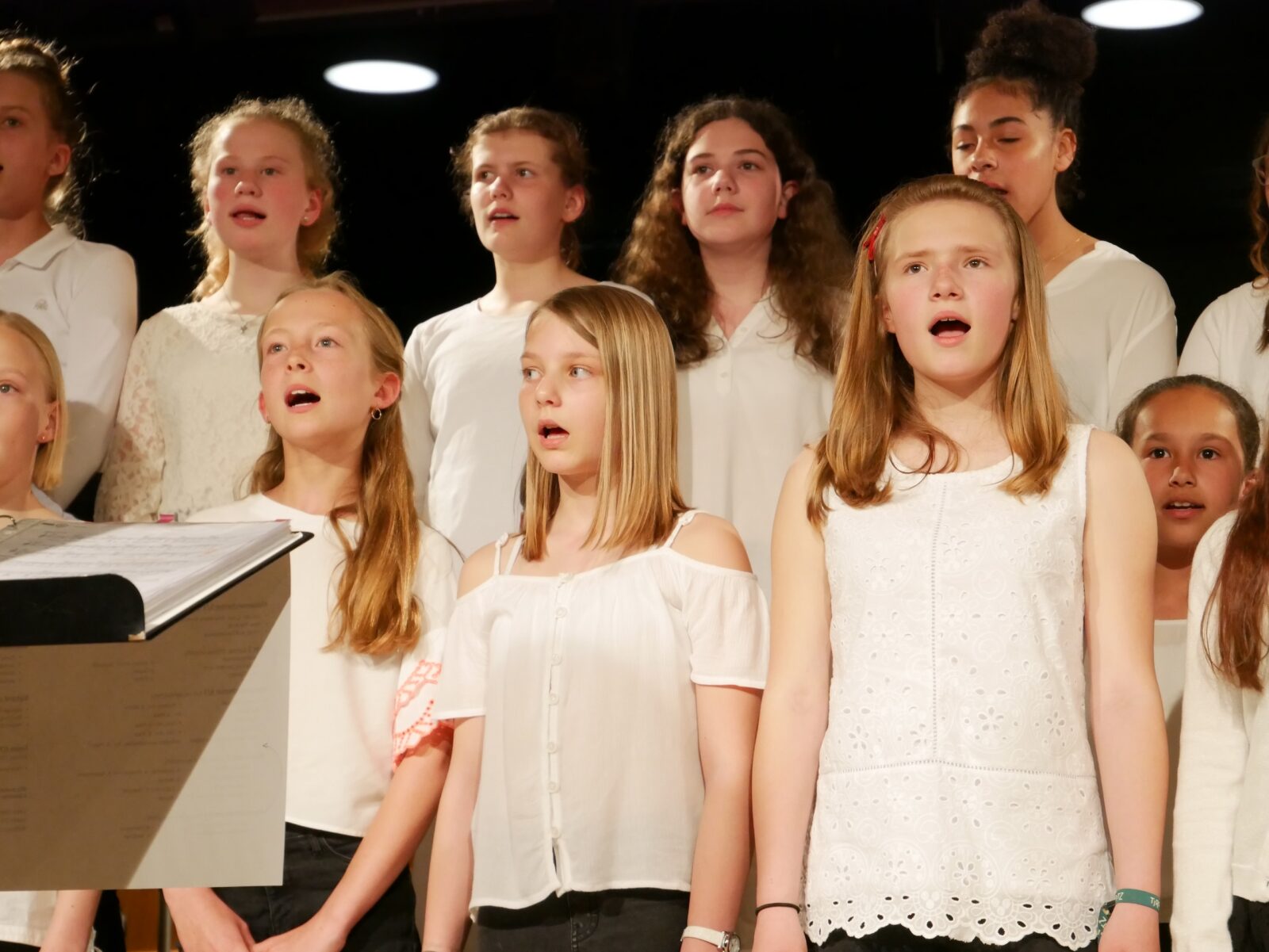 Eine Gruppe von Mädchen in weißen Oberteilen steht gemeinsam auf der Bühne und singt im Chor. Einige haben mitten im Lied den Mund offen und wirken ganz bei der Sache. Im Hintergrund sind der schwarze Hintergrund und die Bühnenbeleuchtung zu sehen.