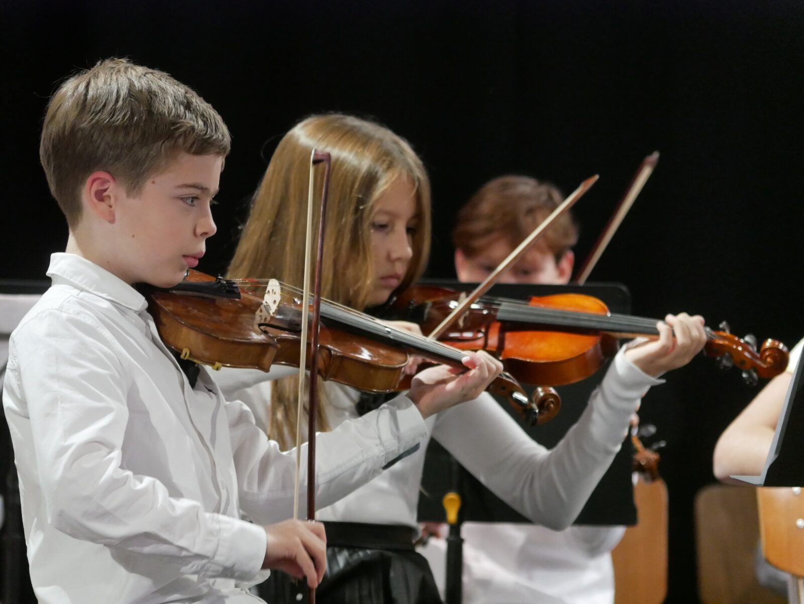 Zwei Kinder in weißen Hemden spielen in einem Orchester Geige und konzentrieren sich ganz auf ihre Musik. Der Hintergrund ist dunkel, hinter ihnen ist ein weiterer Musiker teilweise zu sehen.