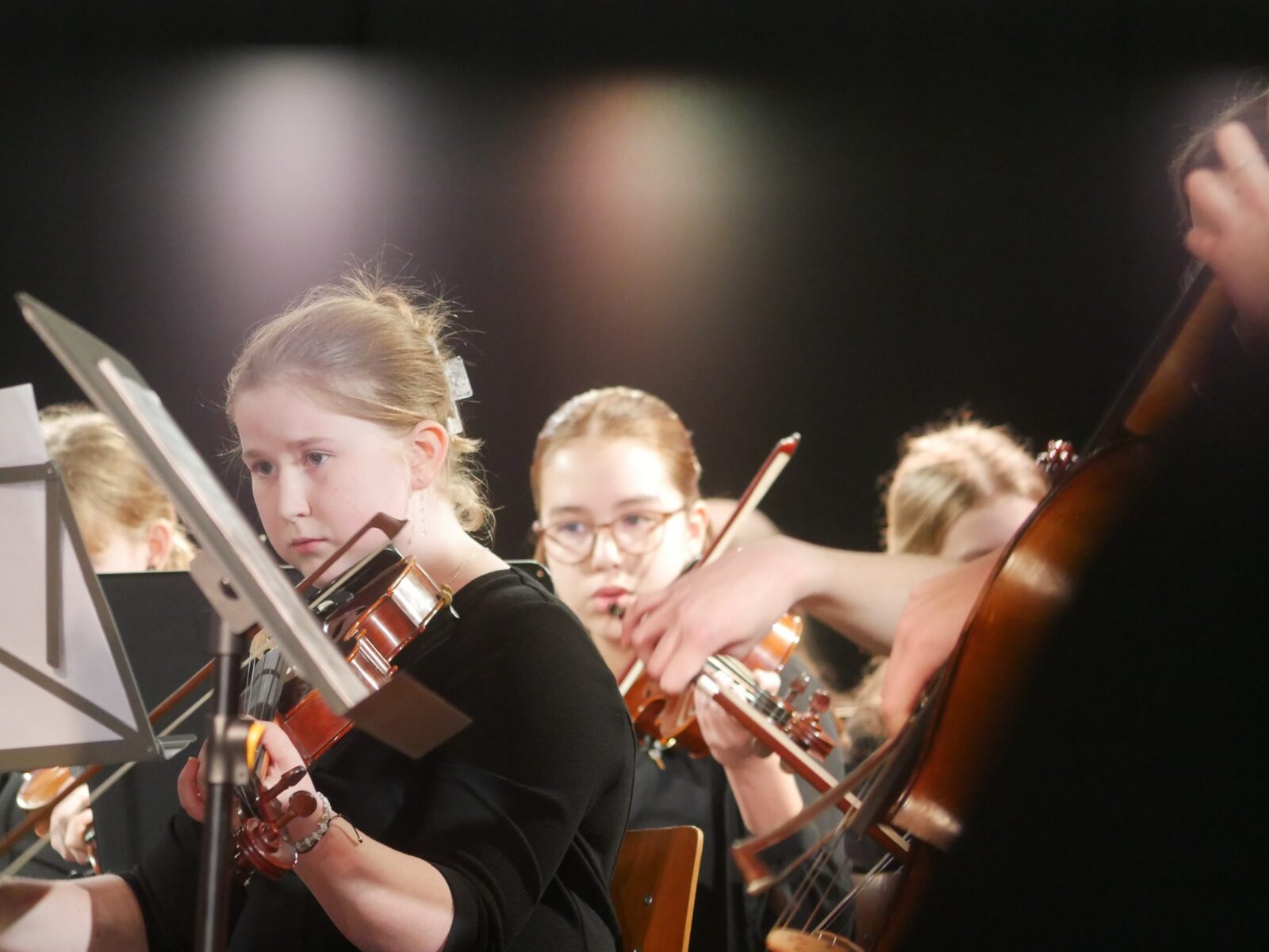 Eine Gruppe junger Musiker spielt unter sanfter Bühnenbeleuchtung Streichinstrumente, darunter Geigen und ein Cello. Im Mittelpunkt steht ein Mädchen im Vordergrund, das während des Spiels Noten liest.