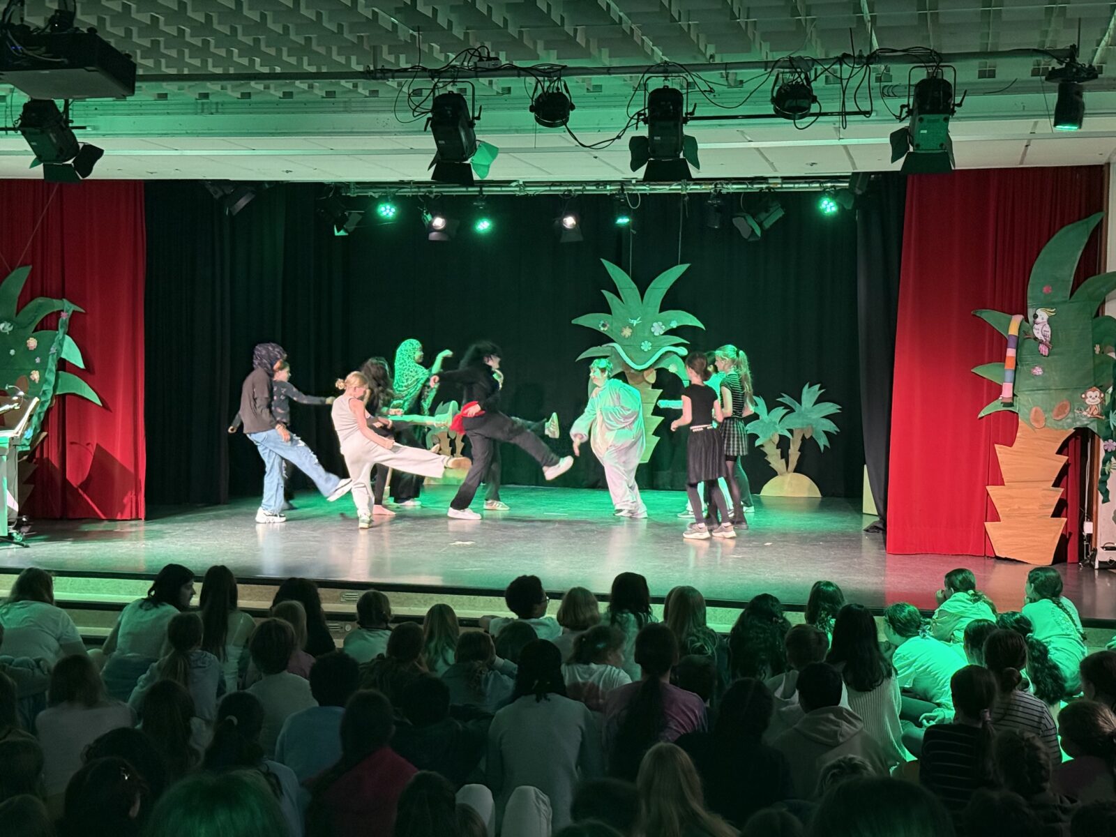 Eine Gruppe von Kindern führt auf einer Bühne mit Dschungel-Dekorationen, darunter Palmen, ein Theaterstück auf, während ein Publikum aus Kindern zusieht. Die Bühne ist grün beleuchtet, und die Darsteller tragen Kostüme.