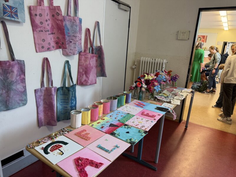 Auf einem Tisch sind farbenfrohe handgefertigte Kunstwerke ausgestellt, darunter verzierte Notizbücher und Stifthalter. An der Wand darüber hängen Batik-Tragetaschen, und auf einem Beistelltisch stehen Kunstblumen. Im Hintergrund haben sich mehrere Personen versammelt.