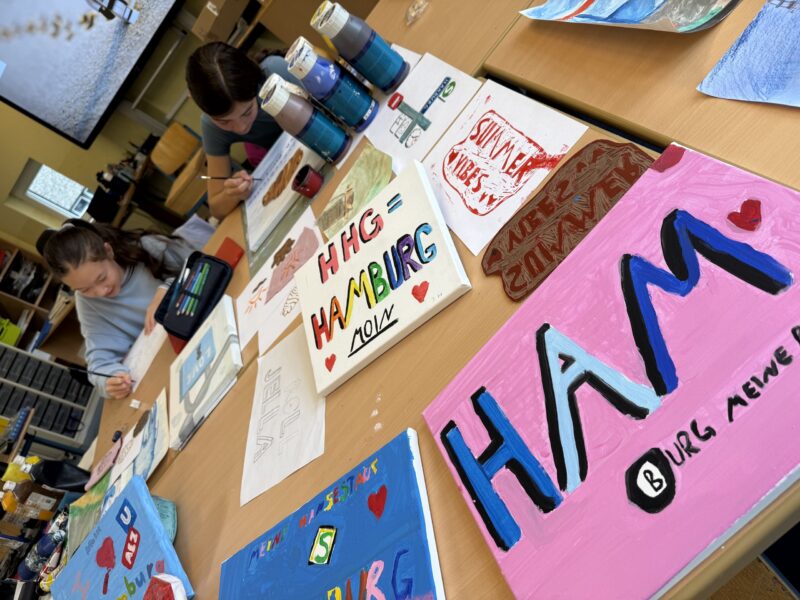 Zwei Kinder sitzen an einem Tisch und gestalten bunte Kunstwerke mit Schildern und Gemälden, auf denen das Wort „HAMBURG“ und farbenfrohe Motive zu sehen sind. Malutensilien, Farbtöpfe und Pinsel liegen verstreut auf dem Tisch.