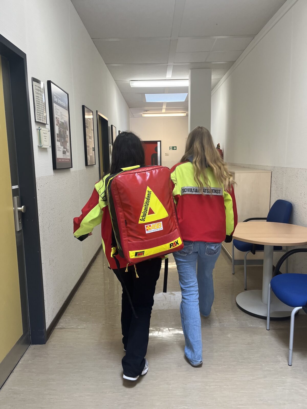 Zwei Personen in roten und gelben Uniformen gehen einen Flur entlang. Eine von ihnen trägt einen großen Notfallrucksack. Beide haben lange Haare und gehen nebeneinander her, wobei einer den Arm um den anderen gelegt hat. Auf der rechten Seite stehen blaue Stühle und ein Tisch.