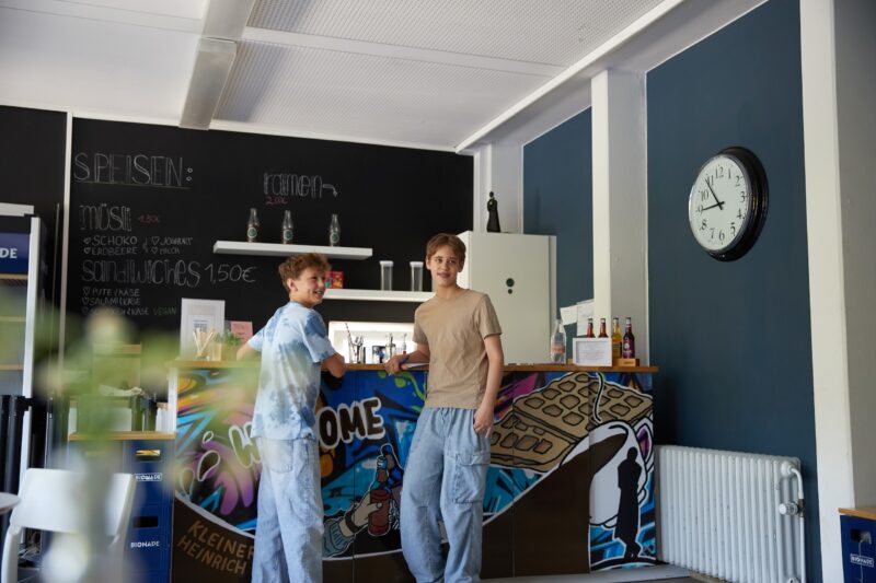Zwei junge Männer stehen lächelnd hinter einer bunten Theke in einem modernen Café mit blauen Wänden, einer Wanduhr und einer Kreidetafel mit der Speisekarte über ihnen. Flaschen und kleine Pflanzen schmücken die Theke.