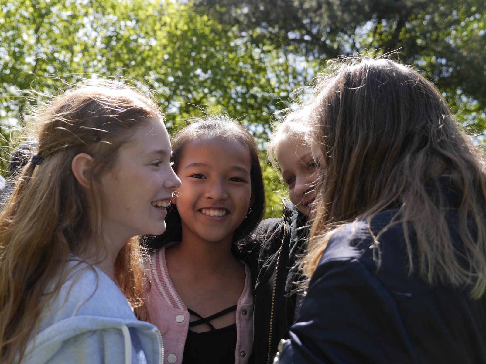 Vier lächelnde Kinder stehen an einem sonnigen Tag im Freien dicht beieinander, im Hintergrund sind grüne Bäume zu sehen. Sie scheinen die Gesellschaft der anderen zu genießen.