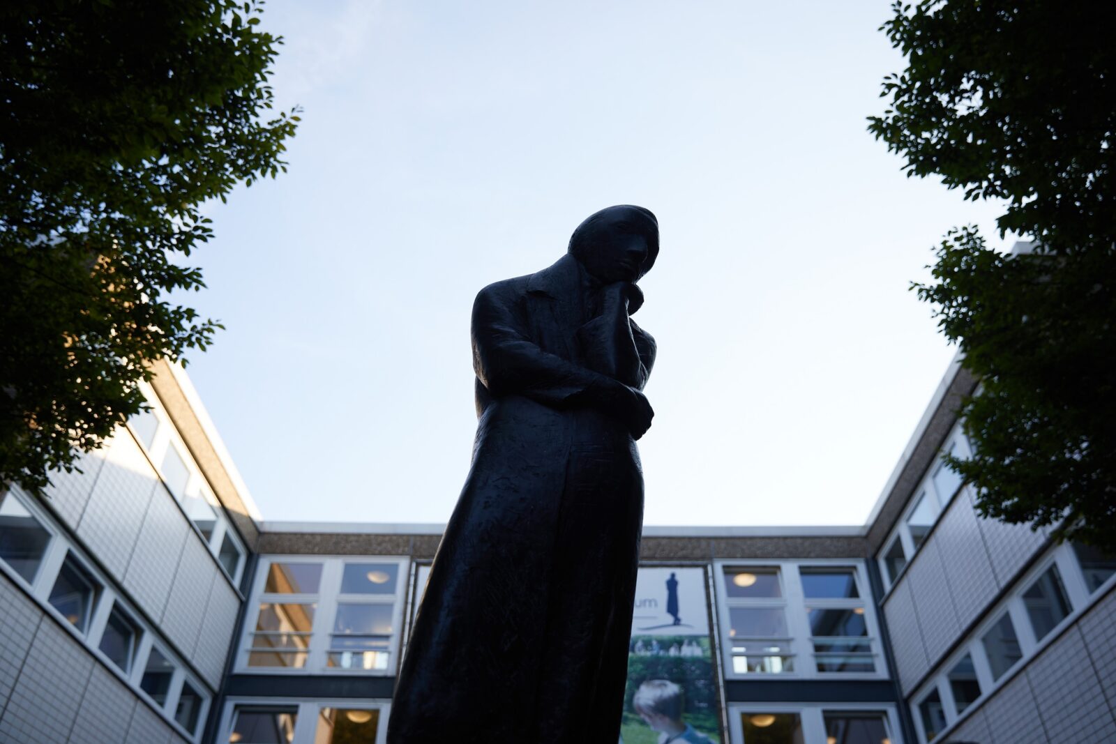 Vor einem modernen Gebäude steht eine dunkle Statue einer Person mit verschränkten Armen, eingerahmt von Bäumen, vor dem Hintergrund eines strahlend blauen Himmels.