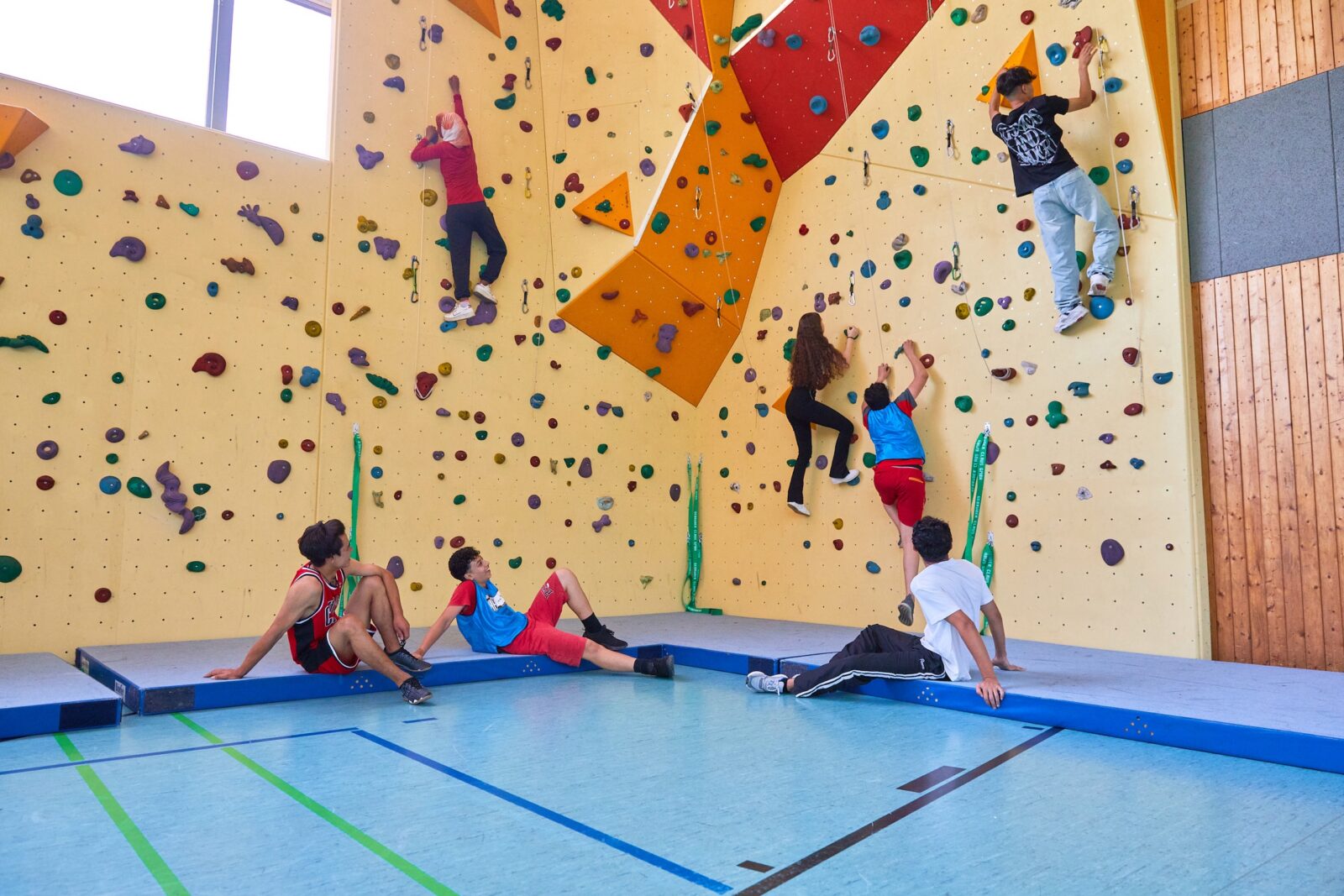 Sechs junge Leute befinden sich in einer Kletterhalle. Drei klettern an einer bunten Boulderwand, während drei andere unten auf blauen Matten sitzen, zuschauen und sich ausruhen. Sonnenlicht fällt durch ein hohes Fenster herein.