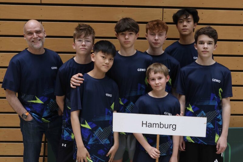 Eine Gruppe aus sieben Jungen und einem Erwachsenen, die alle einheitliche marineblaue Hemden mit geometrischen Mustern tragen, steht gemeinsam in einem Raum. Ein Junge hält ein Schild mit der Aufschrift „Hamburg“ in der Hand. Im Hintergrund ist eine Holzwand zu sehen.