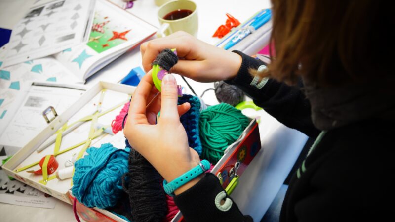 Eine Person mit einem blauen Armband wickelt schwarzes Garn um ein grünes Werkzeug, das auf einem Tisch inmitten von bunten Garnknäueln, einer Schere, Papier und einer Tasse Kaffee liegt.