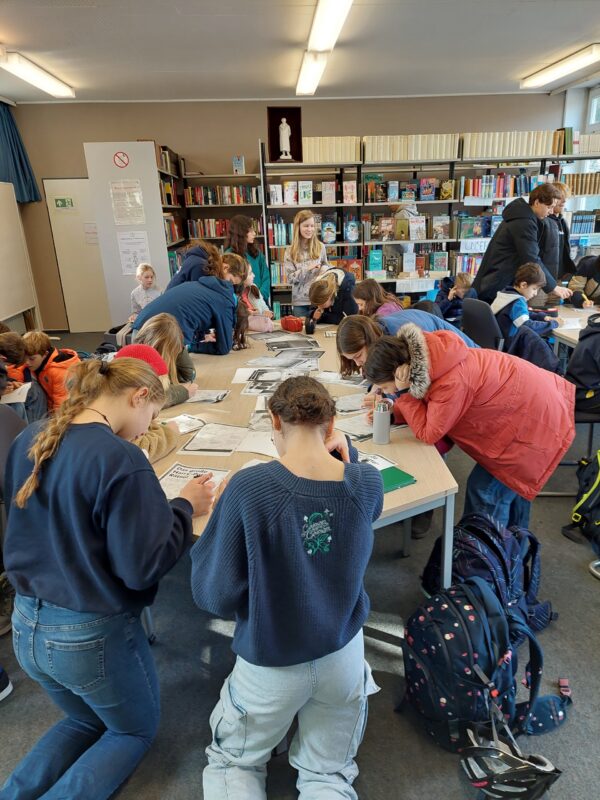 Eine Gruppe von Studierenden arbeitet gemeinsam an Tischen in einer Bibliothek und konzentriert sich auf ihre Unterlagen und Bücher. An den Wänden reihen sich Bücherregale aneinander, und auf dem Boden stehen Rucksäcke. Es herrscht eine geschäftige Atmosphäre, in der alle zusammenarbeiten.