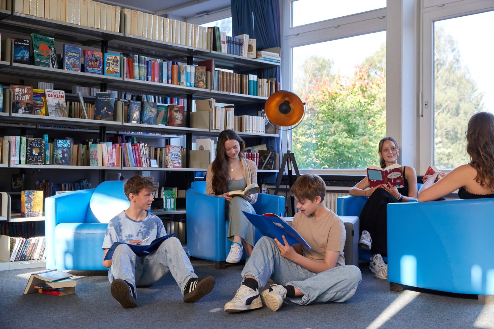 Vier junge Leute lesen in einer Bibliothek Bücher. Zwei sitzen auf blauen Stühlen, während zwei andere auf dem Boden sitzen. Im Hintergrund sind mit Büchern gefüllte Regale und ein großes Fenster zu sehen, durch das Sonnenlicht hereinfällt.