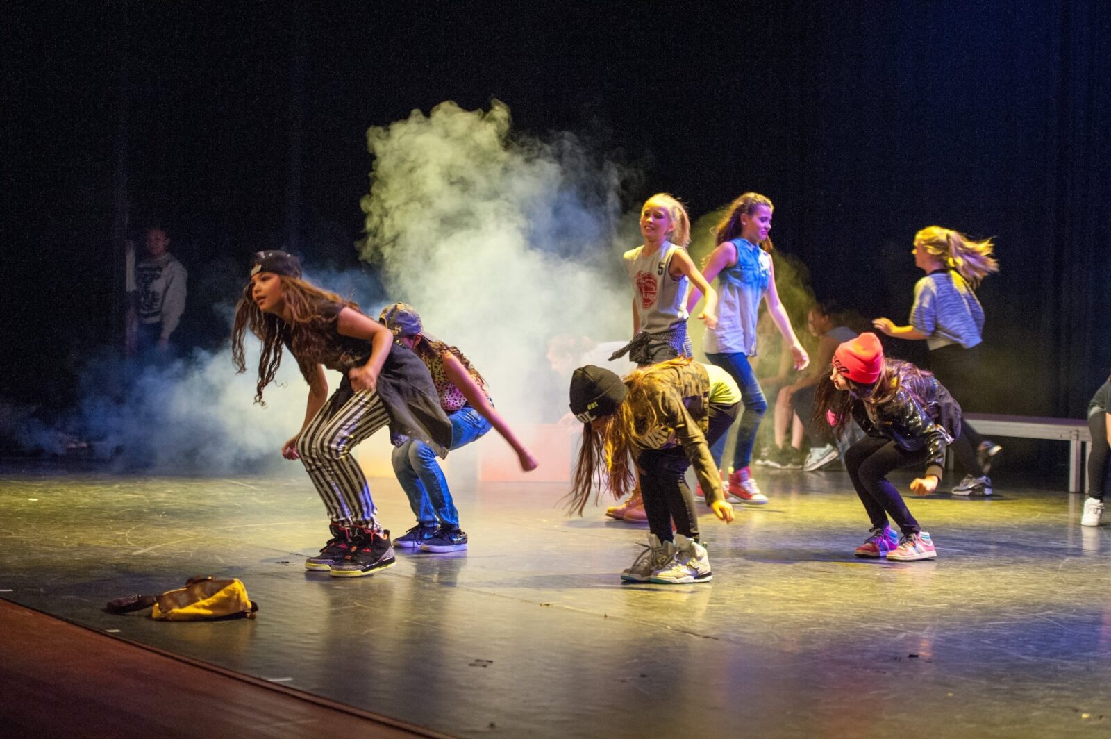 Eine Gruppe von Kindern in bunten, legeren Outfits und Hüten tanzt voller Energie auf einer Bühne mit Raucheffekten und dramatischer Beleuchtung und sorgt so für eine lebhafte Atmosphäre bei der Aufführung.