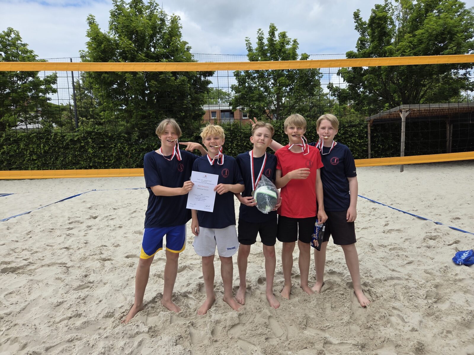 Fünf Jungen stehen auf einem sandigen Volleyballfeld und lächeln mit Medaillen um den Hals. Einer hält eine Urkunde in der Hand, ein anderer einen Volleyball. Sie tragen Sporttrikots und Shorts, im Hintergrund sind Bäume und ein Volleyballnetz zu sehen.