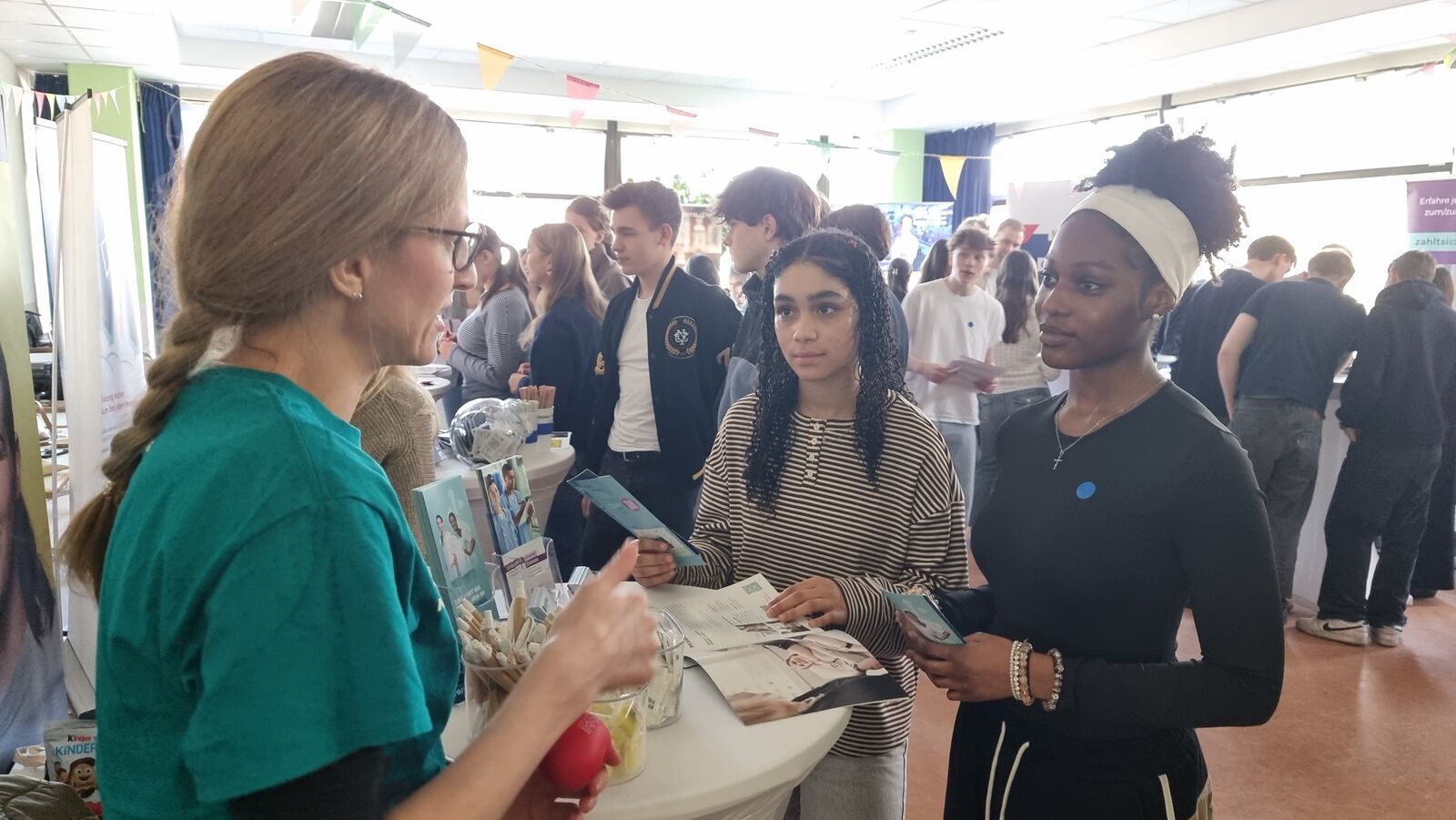 Eine Frau in einem blaugrünen Shirt unterhält sich bei einer gut besuchten Indoor-Veranstaltung mit zwei jungen Frauen. Sie halten Faltblätter in der Hand und stehen neben einem runden Tisch, auf dem Broschüren und Kleinigkeiten ausliegen. Im Hintergrund sind weitere Personen und Stände zu sehen.