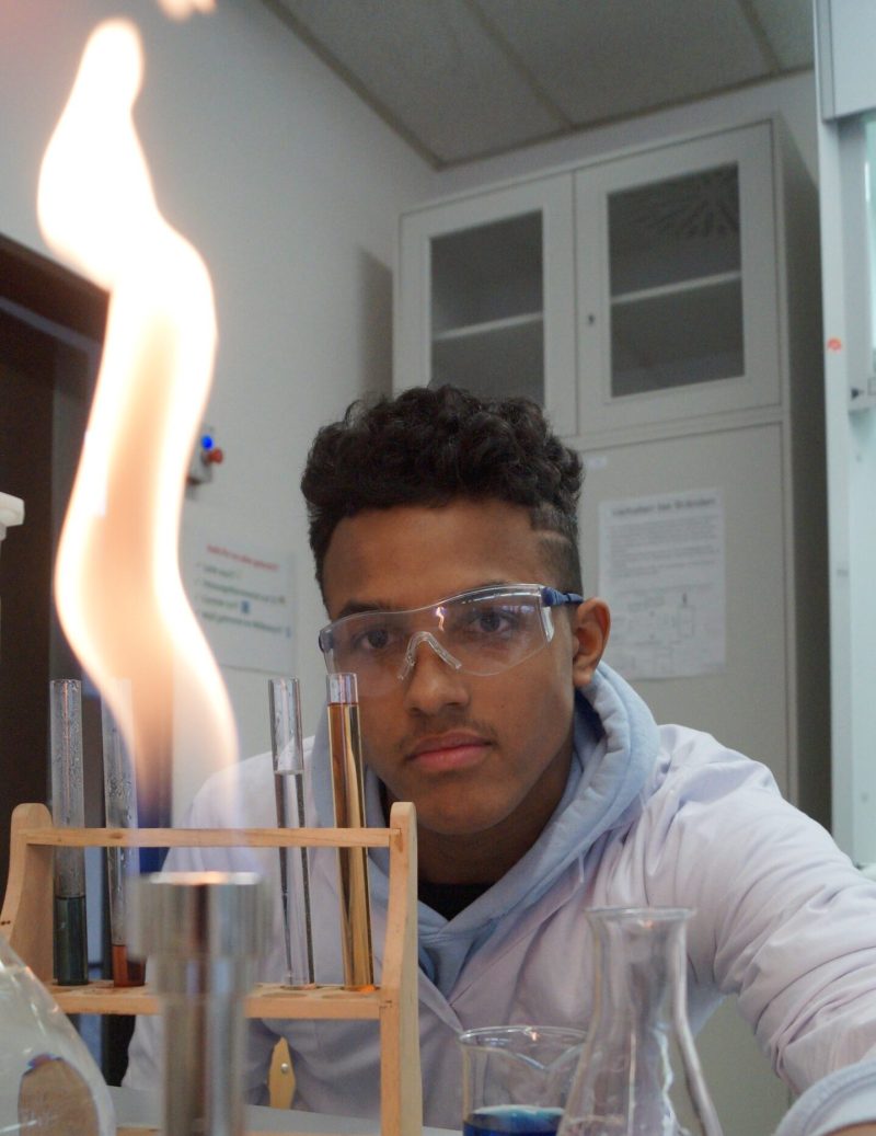 Ein Sch&uuml;ler, der eine Schutzbrille und einen Laborkittel tr&auml;gt, beobachtet in einem Chemielabor eine hohe orangefarbene Flamme eines Bunsenbrenners, w&auml;hrend in der N&auml;he Reagenzgl&auml;ser und Kolben mit bunten Fl&uuml;ssigkeiten stehen.